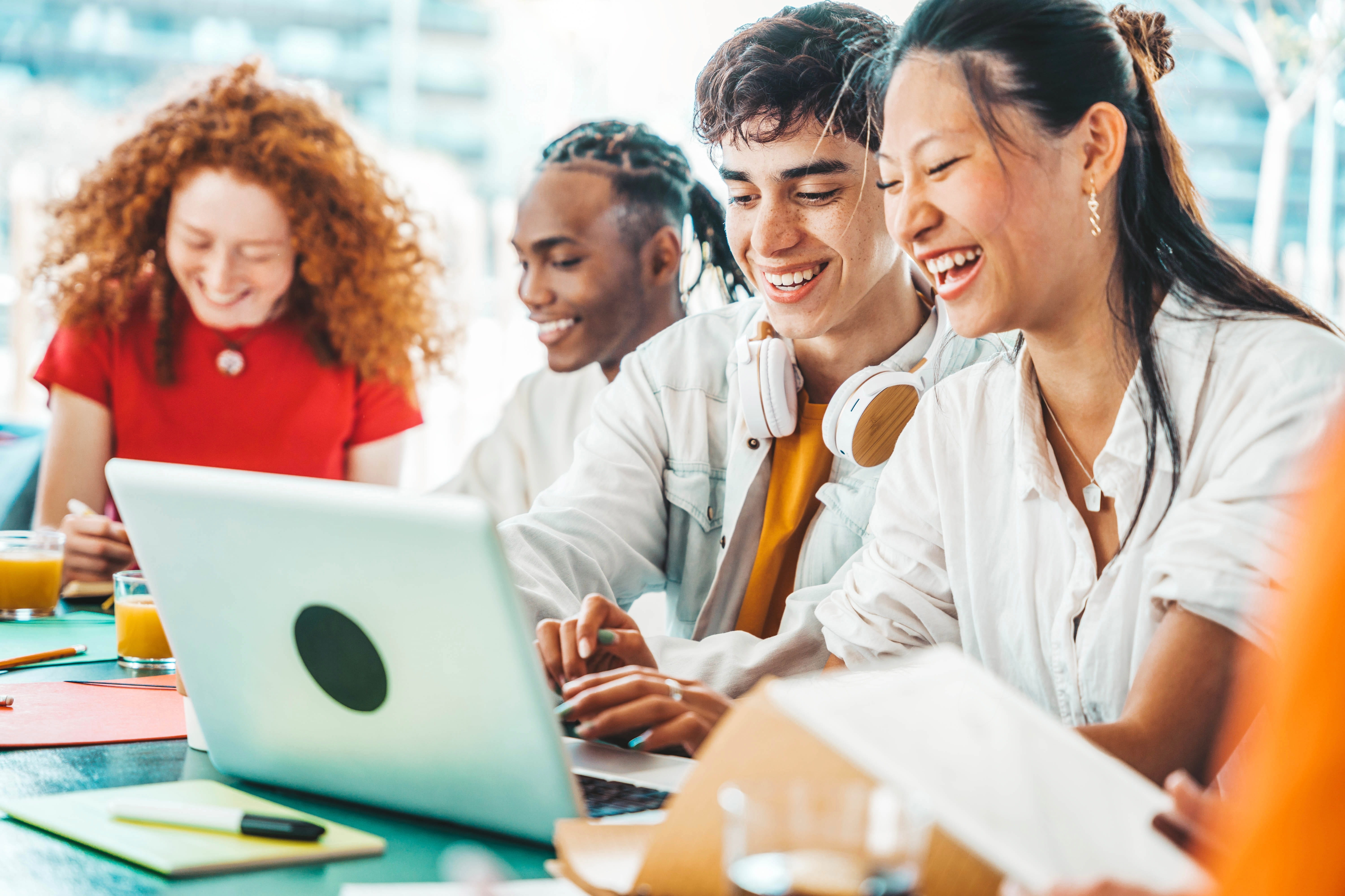 A diverse group of young people collaborating and smiling while using a laptop at a bright, social workspace.