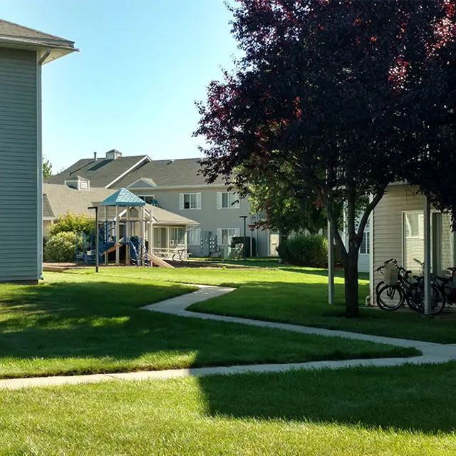 Apartment Complex Playground A grassy area in an apartment complex featuring two buildings, a children's playground with slides and swings, and parked bicycles under a tree.