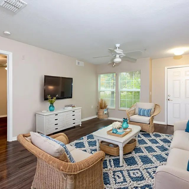 A cozy living room featuring a beige sofa adorned with blue pillows, a light wooden coffee table, and a stylish rug. There is a television mounted on the wall, and large windows allow natural light to fill the space.