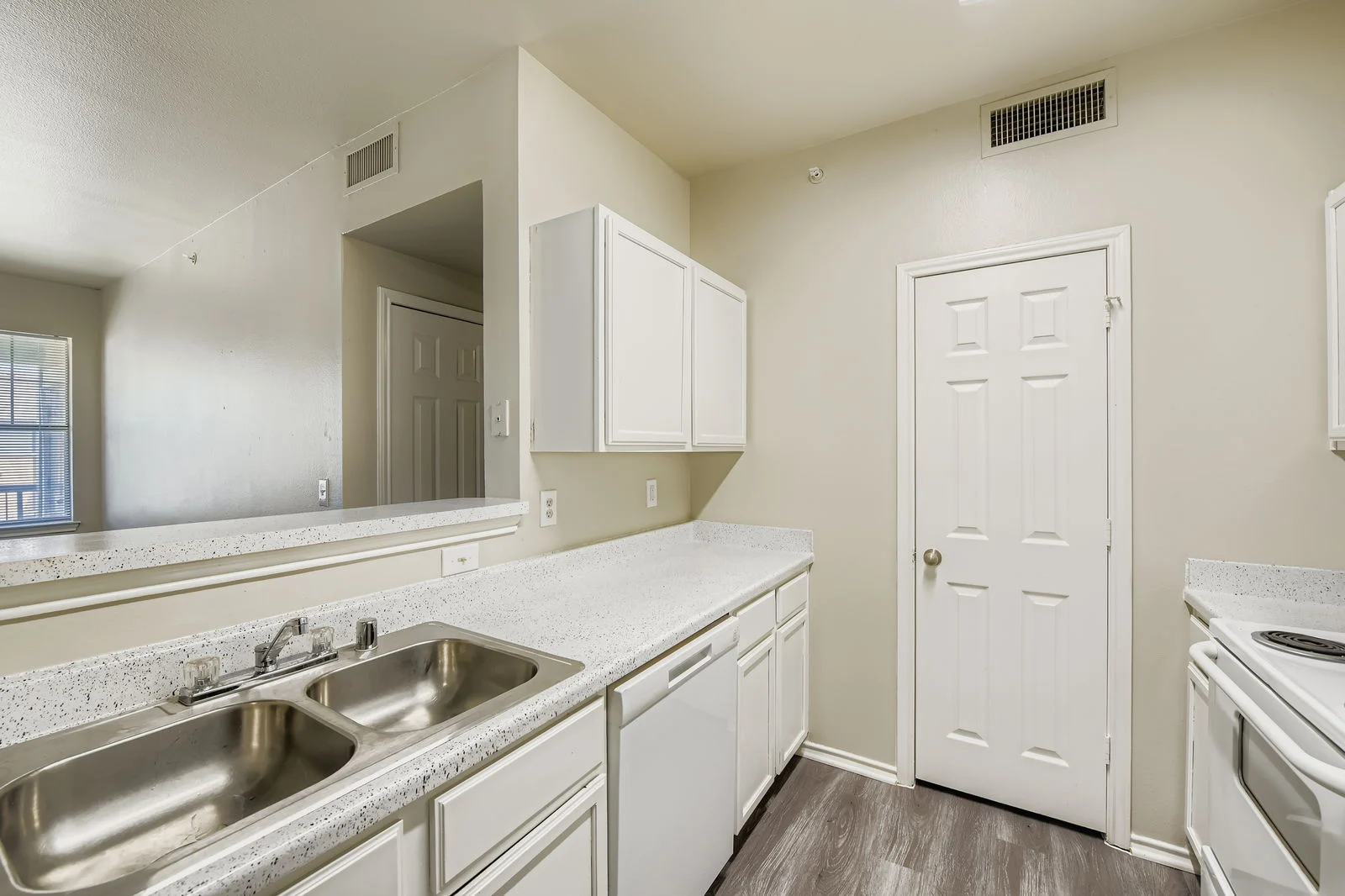 A modern kitchen featuring white cabinetry, a double sink, a countertop with speckled design, and a white door leading to another room. Natural light enters through a window on the left.