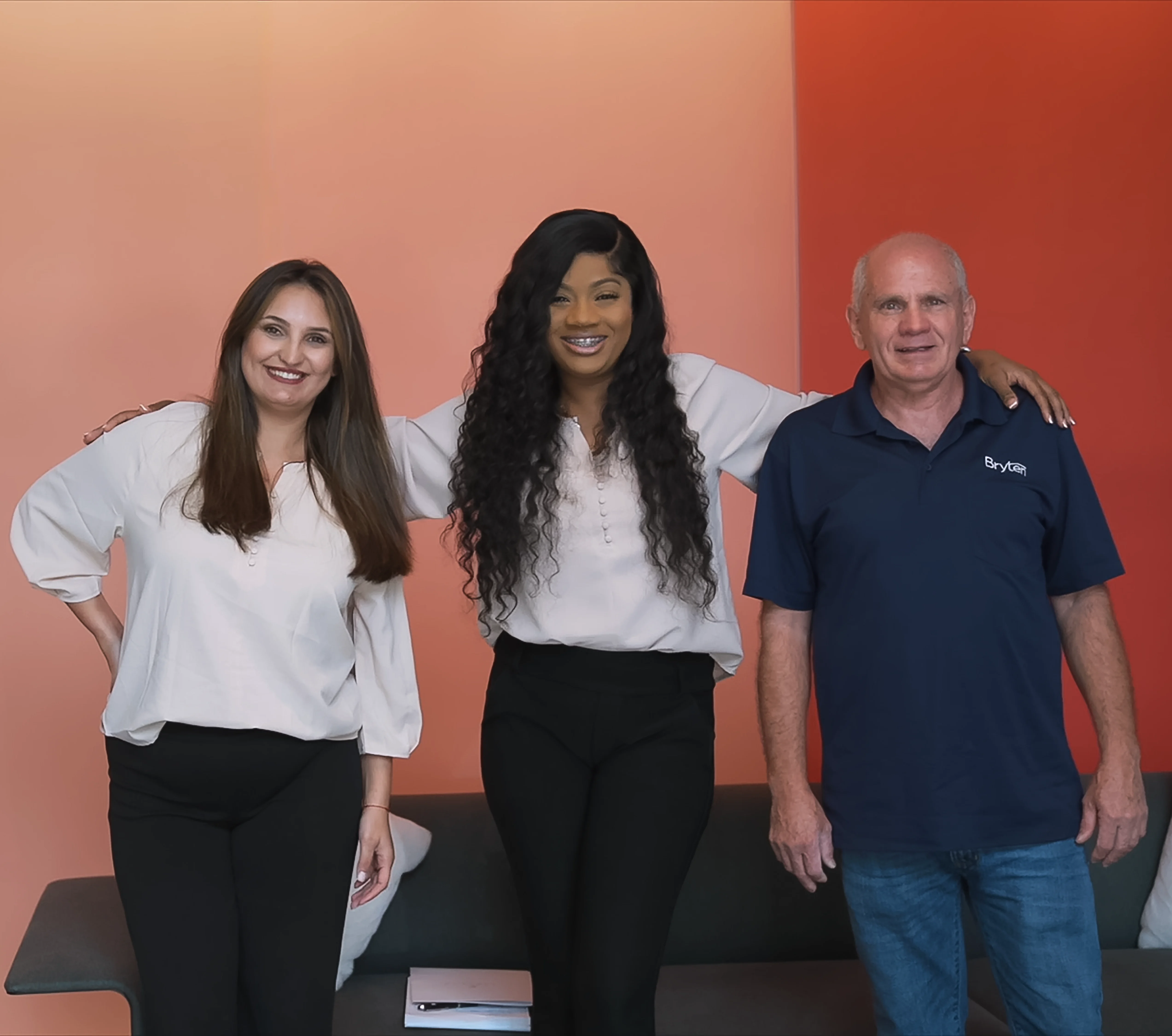 Three people standing together in an office setting, smiling at the camera. Two women and one man, dressed in professional attire, with an orange wall in the background.