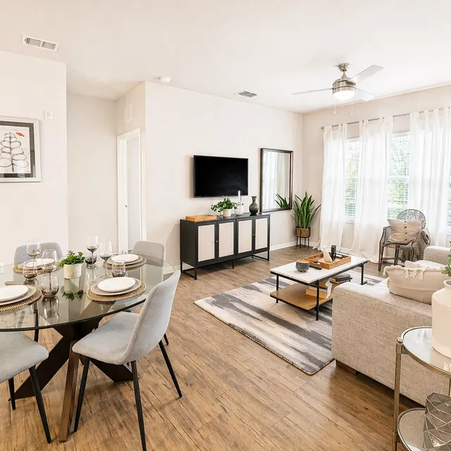 A modern living room and dining area featuring a stylish table set for four, a comfortable sofa, a TV unit, and large windows with natural light.