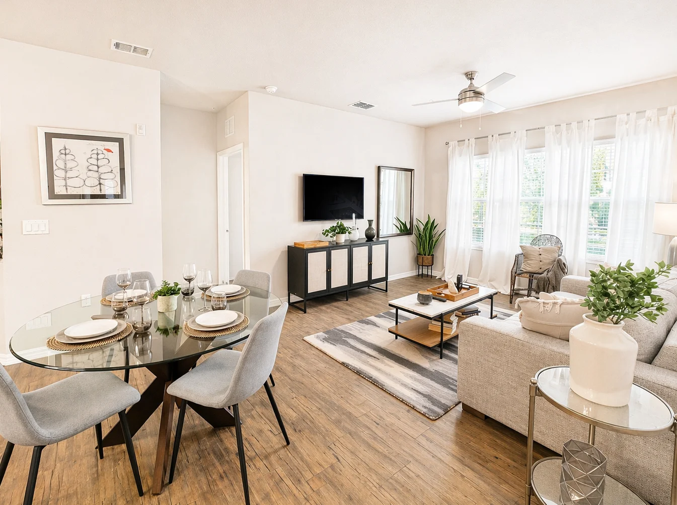 A modern living room and dining area featuring a stylish table set for four, a comfortable sofa, a TV unit, and large windows with natural light.