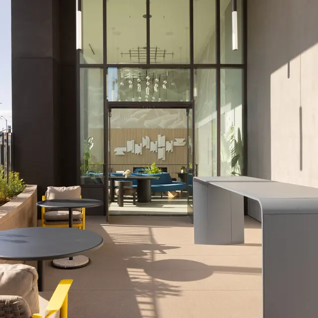 A modern outdoor lounge area featuring sleek furniture, a long gray table, round blue tables, and comfortable seating. Lush greenery is present alongside the seating, and large windows lead into a well-lit indoor space.