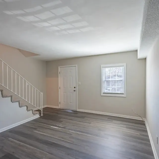 An empty living room with gray walls and dark wood flooring. A staircase is seen on the left side, leading to an upper level. A white door and a window are visible on the opposite wall, allowing natural light to enter the space.
