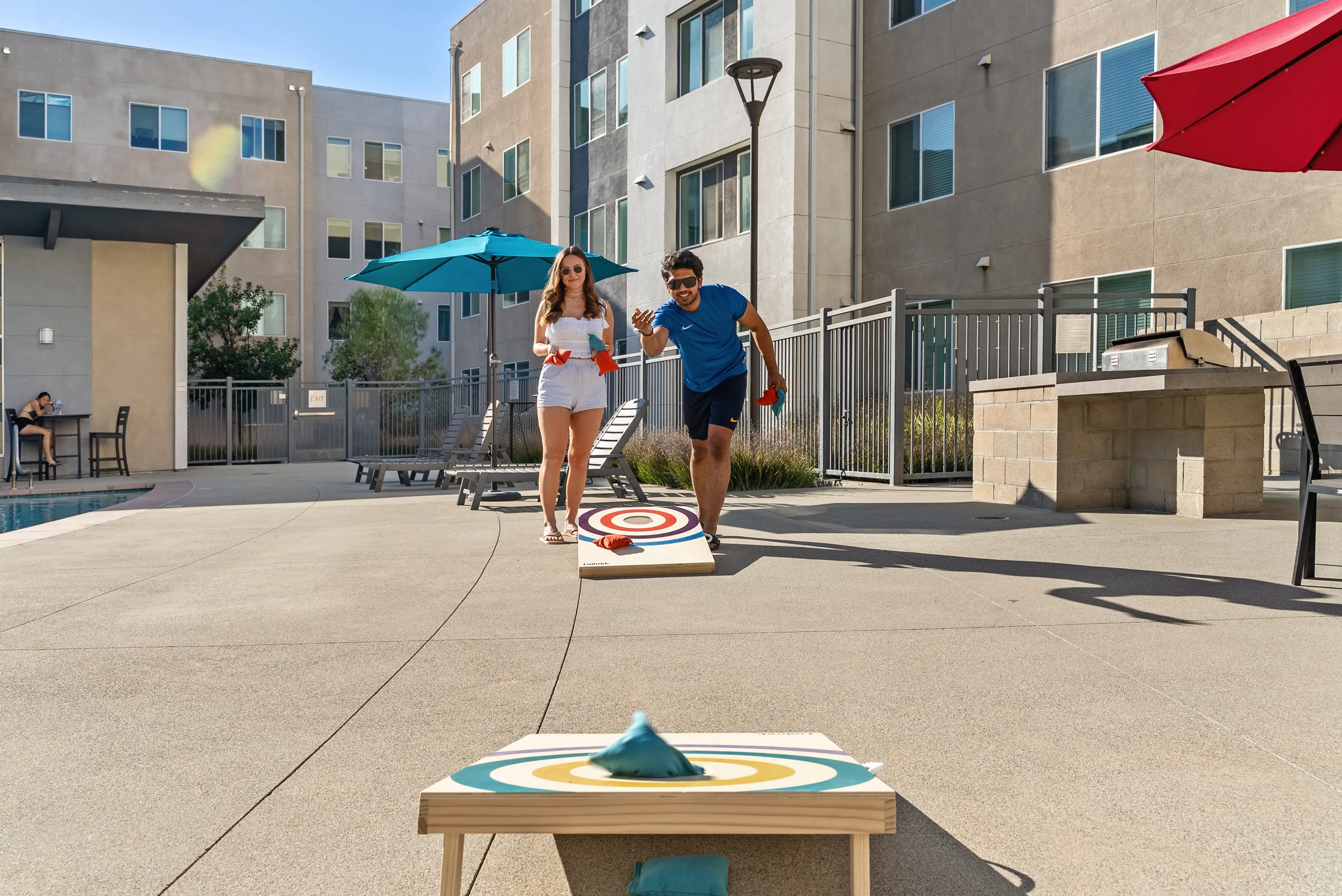 Poolside Cornhole Game A man and woman playing cornhole outdoors by a pool. The woman is wearing a white outfit and the man is in a black shirt with sunglasses, both are poised to throw bean bags towards a cornhole board. There are umbrellas providing shade and the background includes a pool and apartment buildings.