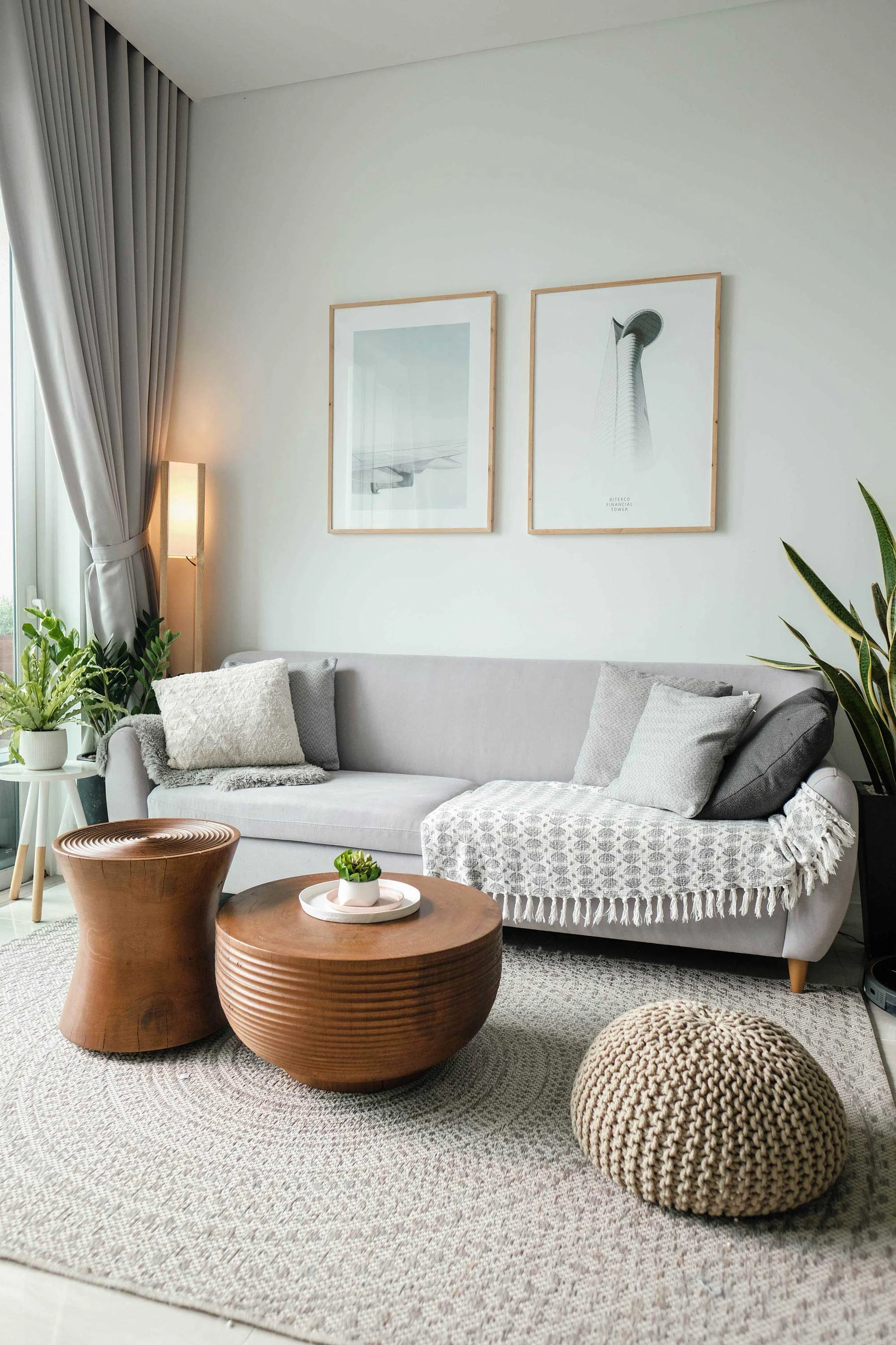 Modern Living Room Design A modern living room featuring a gray couch adorned with a decorative blanket, round wooden coffee table with a small plant, a knitted ottoman, and framed wall art. Large windows with curtains allow natural light, and greenery is visible nearby.