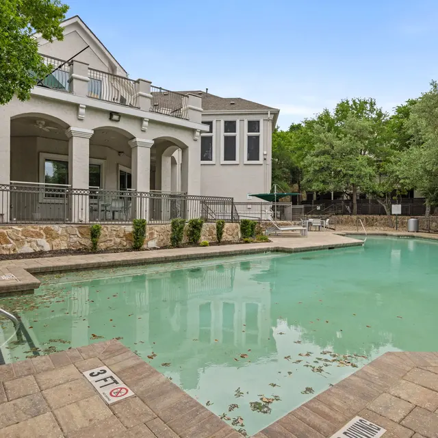 An outdoor swimming pool surrounded by a stone deck and well-maintained landscaping. The pool is partially covered with leaves and features a shallow end with a sign indicating depth. A building with multiple windows is visible in the background, along with trees providing shade.