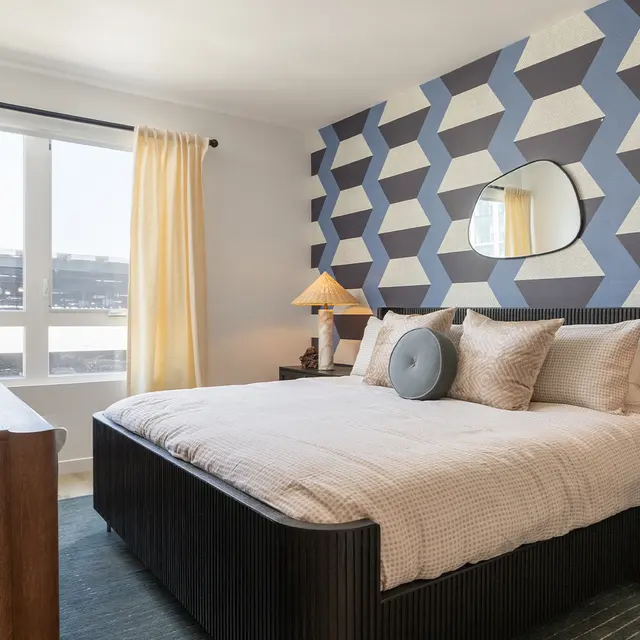A modern bedroom featuring a geometric patterned accent wall, large windows with sheer curtains, a bed with decorative pillows, and stylish lamps on nightstands.