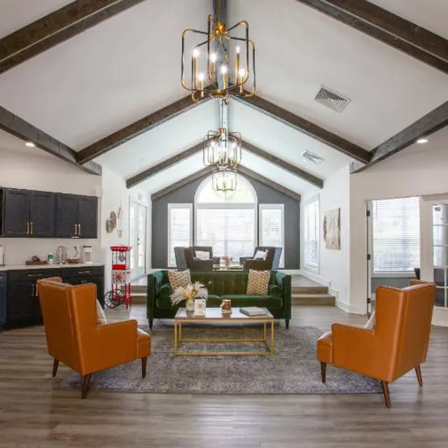 A spacious modern living room featuring vaulted ceilings with wooden beams, two orange armchairs, a green sofa, a coffee table, and a well-equipped kitchen area with black cabinets and stylish lighting.