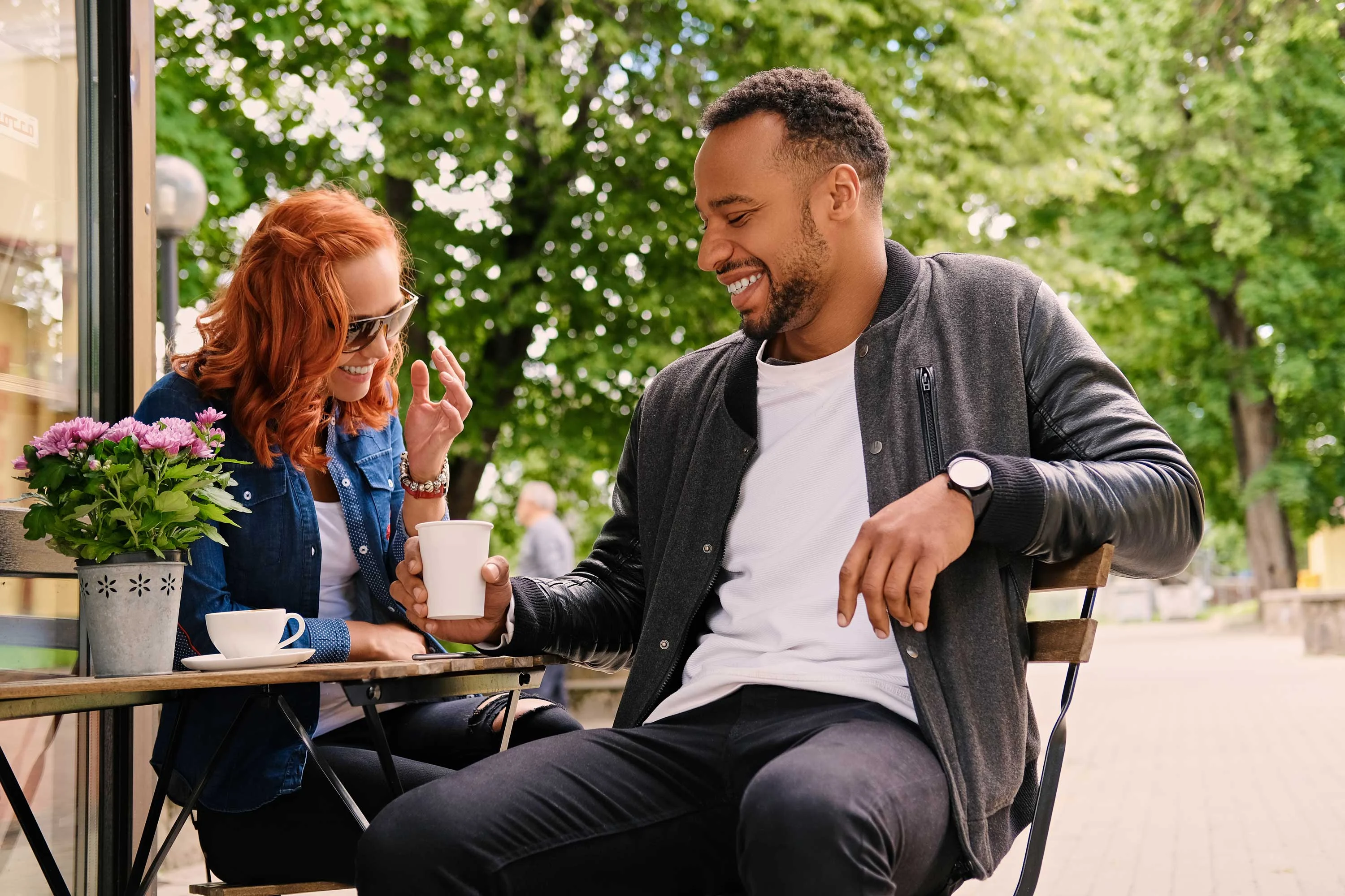 Couple Enjoying Coffee at Café A happy couple sitting at an outdoor café table, laughing and enjoying coffee. The woman has long red hair and sunglasses, while the man is wearing a black and gray jacket. A small potted plant with purple flowers is on the table.