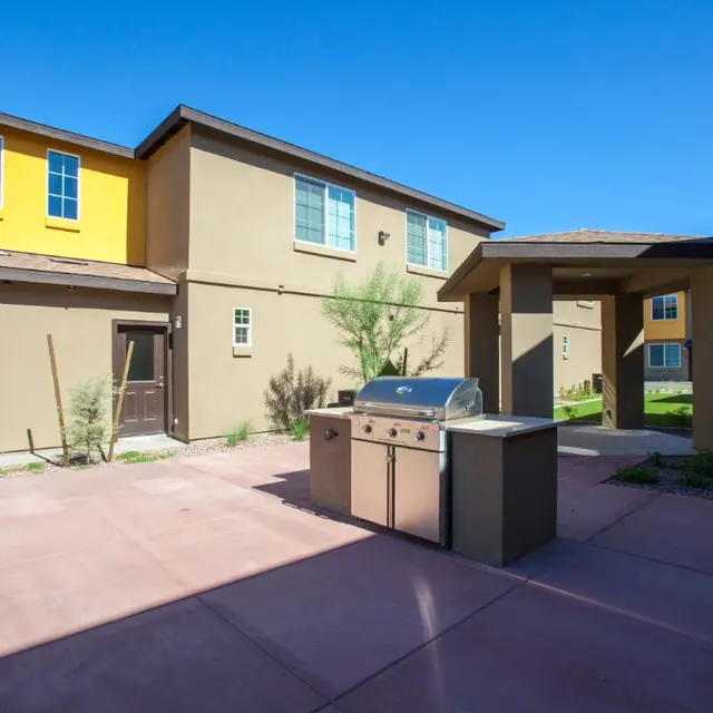 A sunny backyard area featuring a stainless steel grill, with two residential buildings in the background, one with a yellow accent and green landscaping.