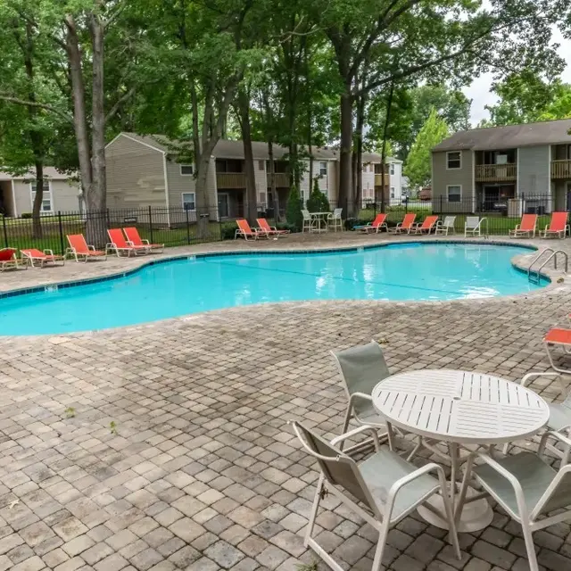 A swimming pool surrounded by lounge chairs and tables in a landscaped area with trees and residential buildings in the background.