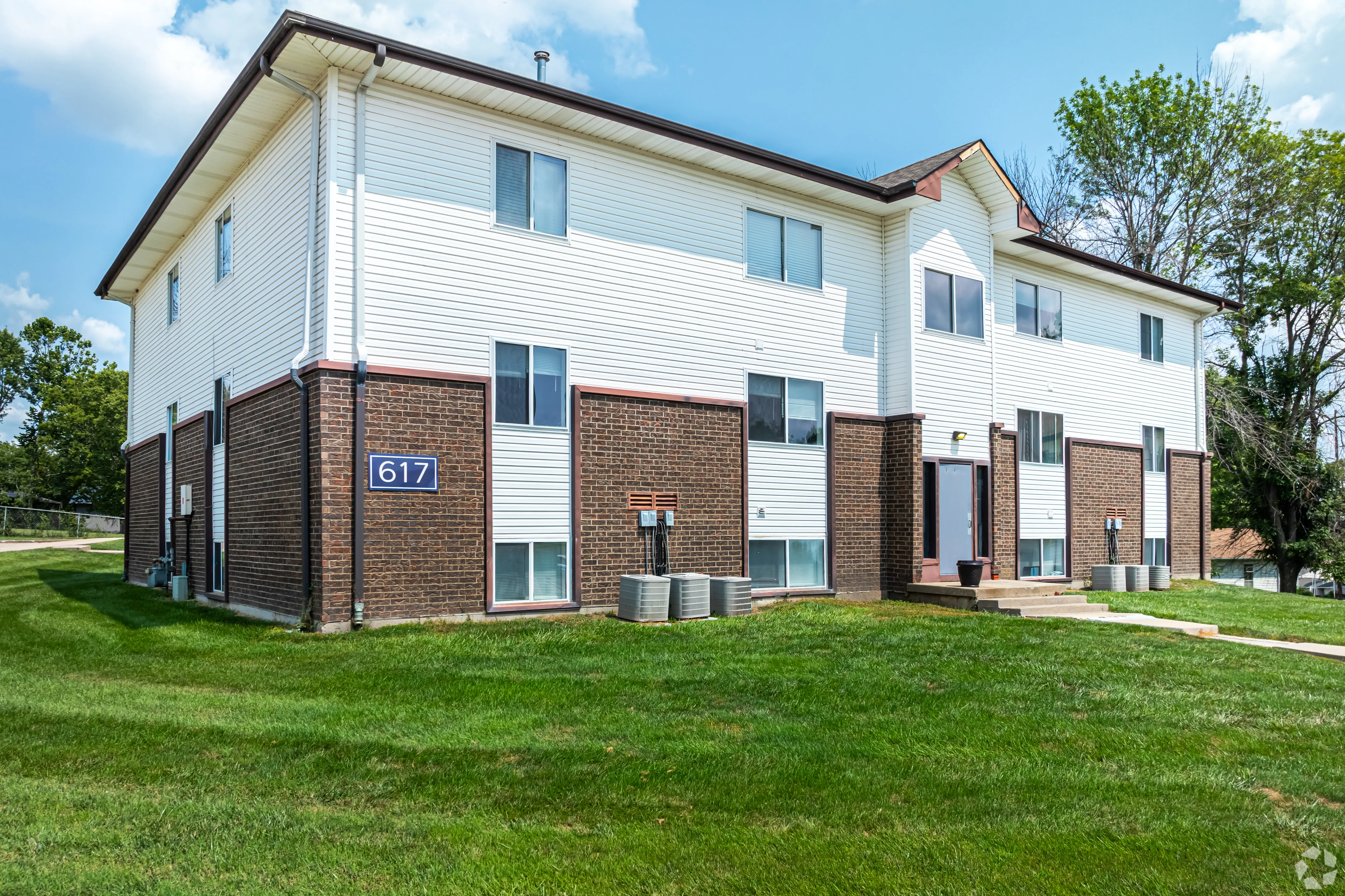 A two-story apartment building with a combination of brick and siding. The building features several windows, a blue door, and a visible address number 617. Green grass surrounds the building, and there are several air conditioning units visible outside. The sky is partly cloudy, giving a warm look to the scene.
