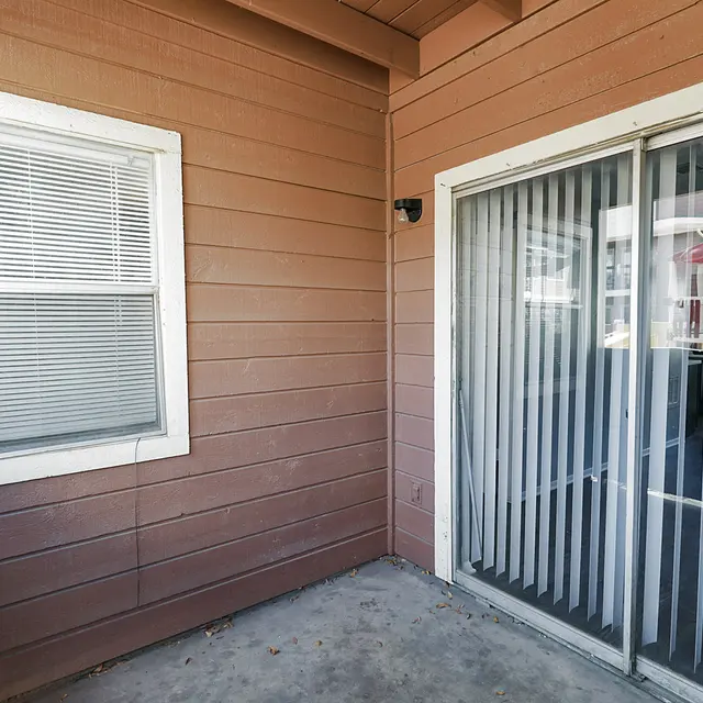 A corner of a patio featuring a window and sliding glass door with vertical blinds, surrounded by brown wooden walls.