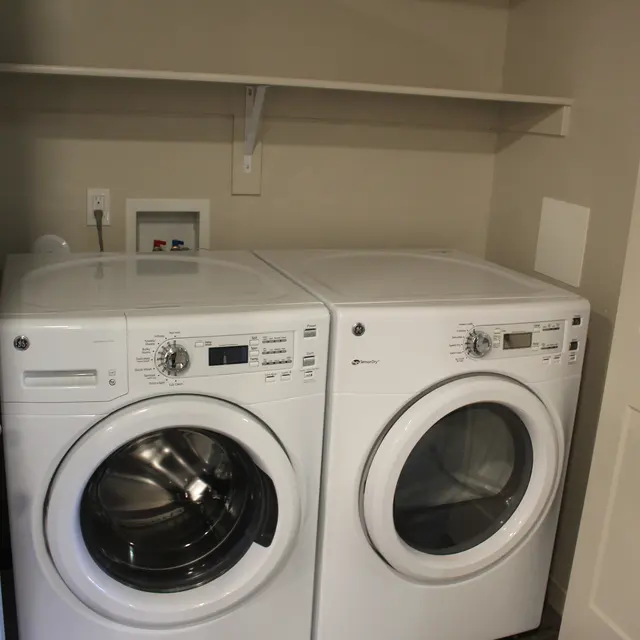 A compact laundry room featuring a stacked washer and dryer set in white, with a shelf above and a door partially visible on the right.