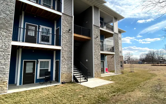 A modern three-story apartment building with a combination of stone and wood siding. The building features balconies, a central staircase, and is surrounded by green grass and a clear blue sky with fluffy clouds.