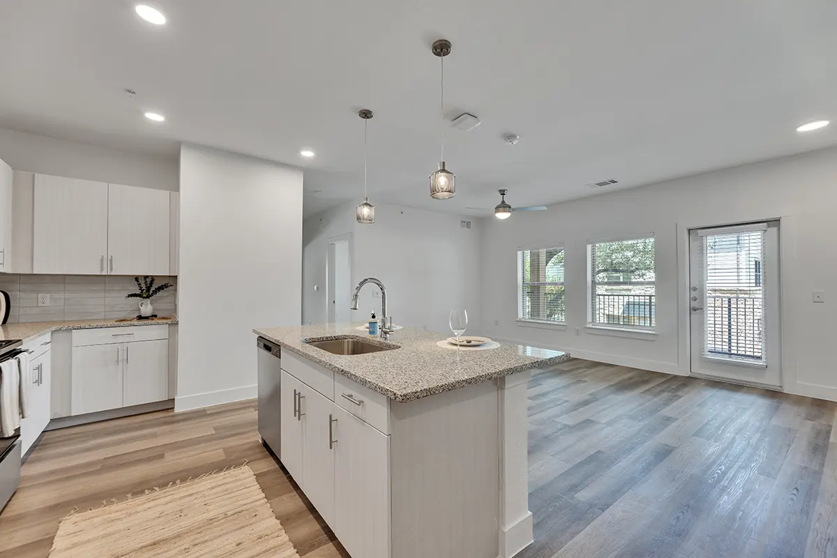 Interior view of a modern apartment featuring a kitchen with white cabinets, a granite island, and a spacious living area with large windows.