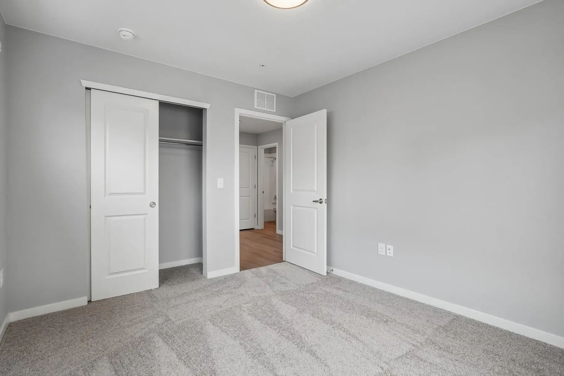 Empty Bedroom Space A spacious empty bedroom with gray walls and carpet. Two open doors lead to closets, while a bright light fixture illuminates the room.
