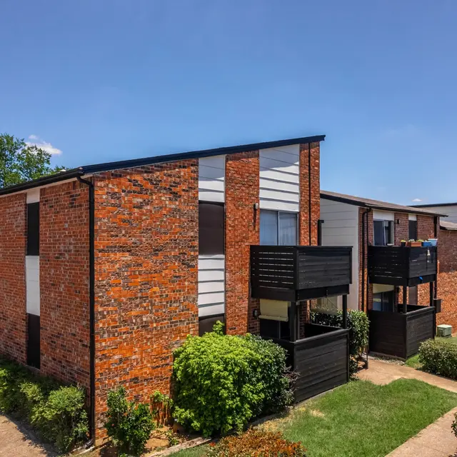 Exterior view of a two-story apartment complex with brick walls and balconies, surrounded by well-maintained landscaping under a blue sky.