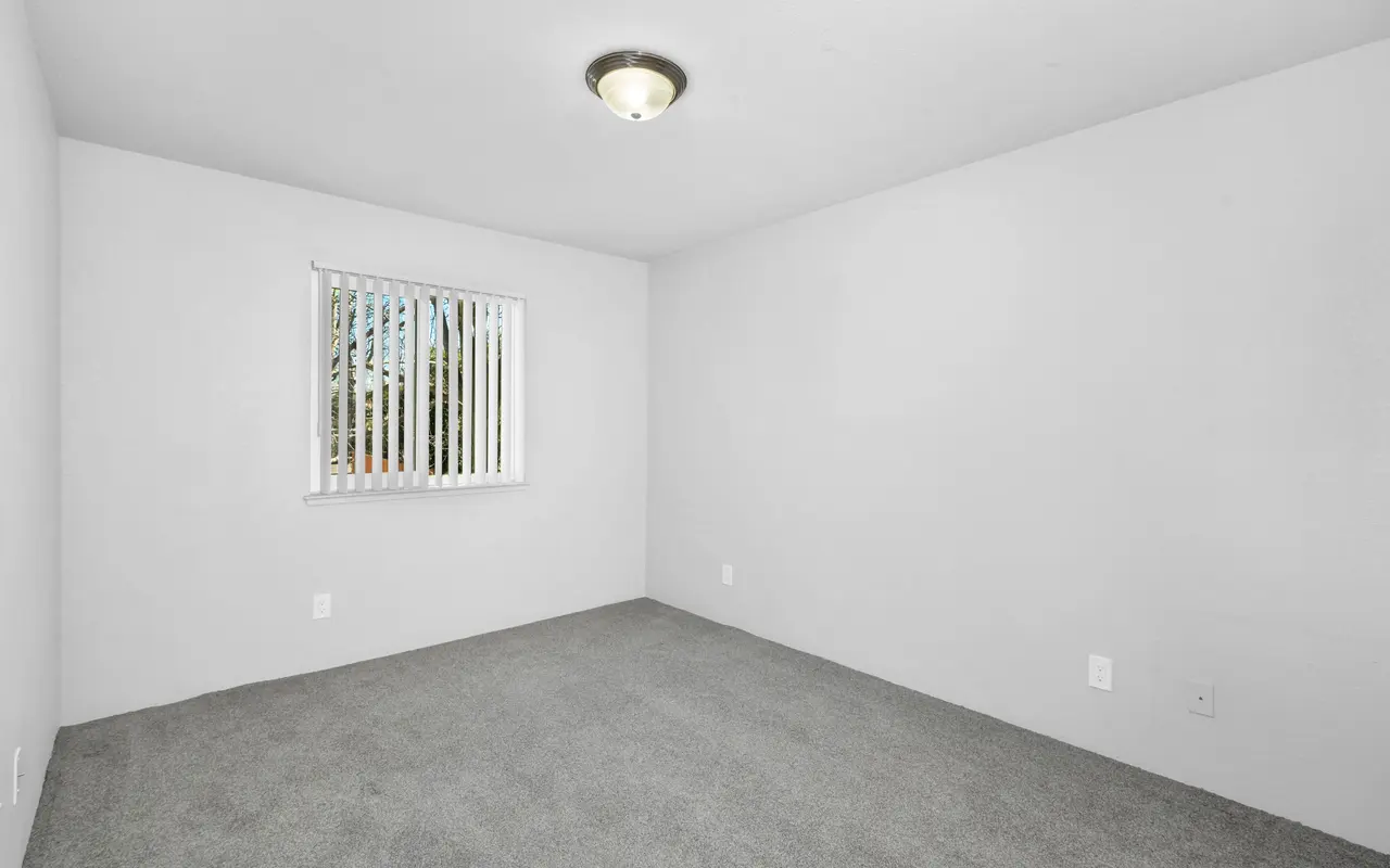 A vacant room with light gray walls and a gray carpet. It features a window with vertical blinds letting in natural light and a ceiling light fixture. The overall atmosphere is minimalistic and uncluttered.