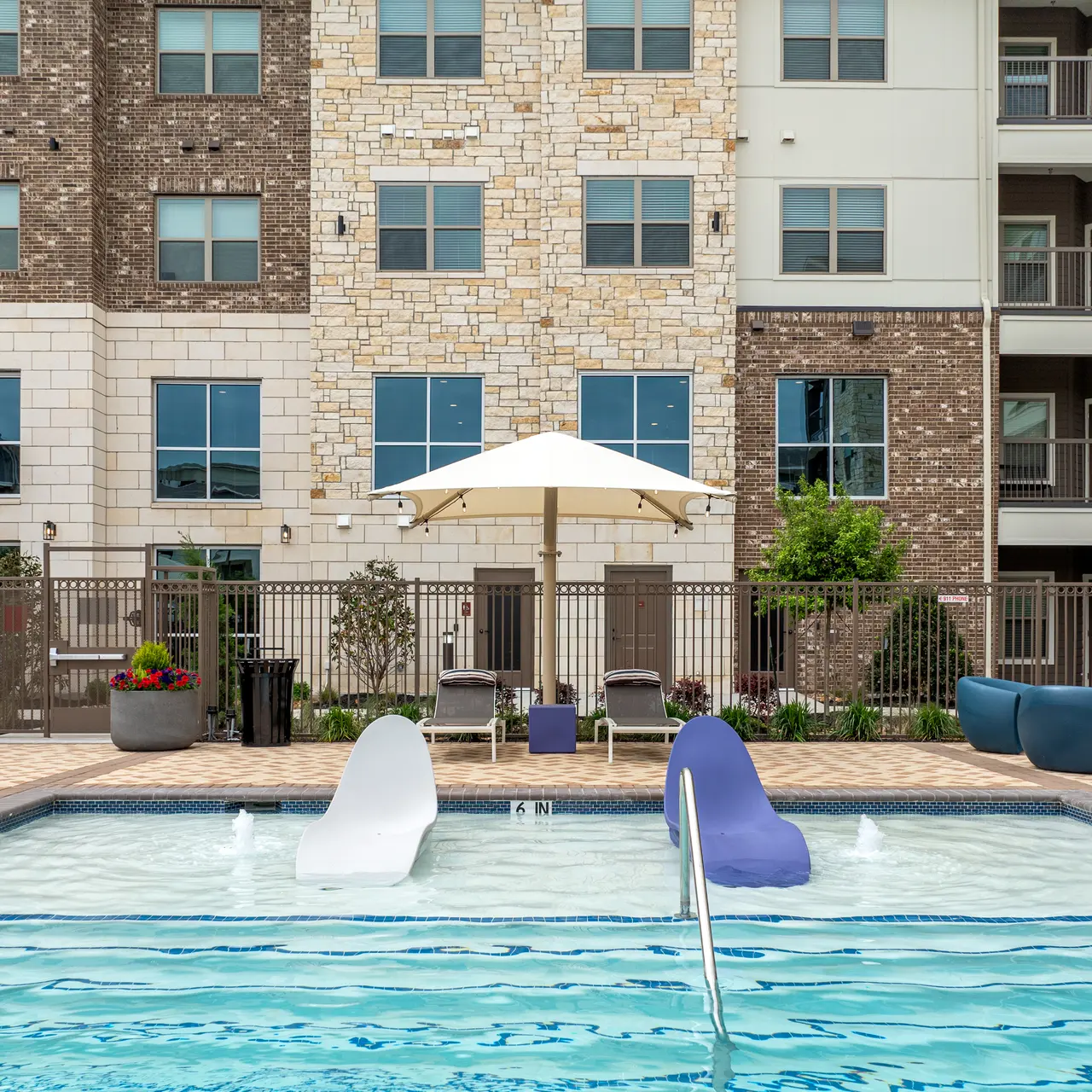 Apartment Pool Area A modern apartment pool area featuring a swimming pool with decorative fountains, surrounded by comfortable lounge chairs and greenery. An umbrella shades seating areas near the pool.