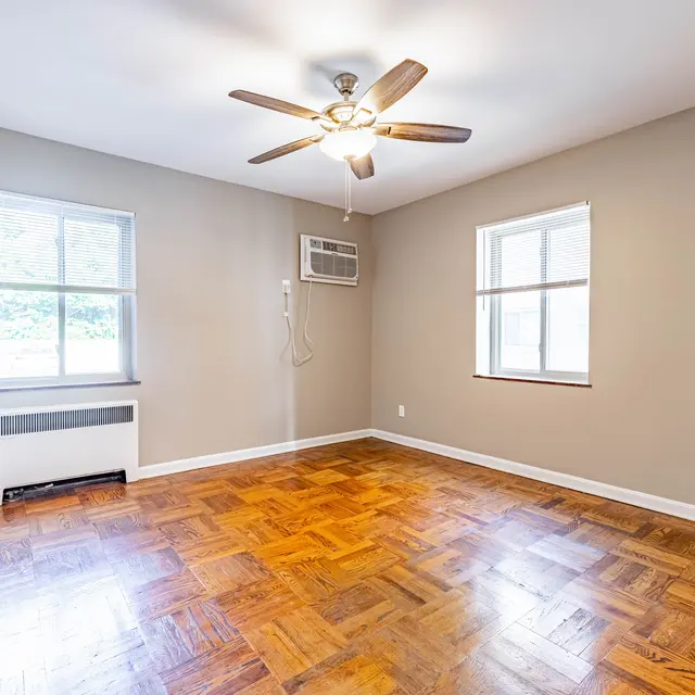 An empty living room featuring wooden flooring, two windows, an air conditioning unit, and a ceiling fan.