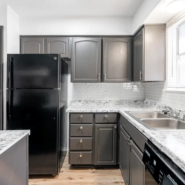A modern kitchen featuring dark cabinets, a black refrigerator, and white granite countertops. The layout includes a double sink with a window above and a small island area with a stove.