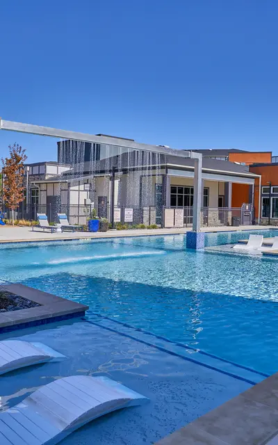 A spacious modern pool area surrounded by sun loungers and apartment buildings, under a clear blue sky.
