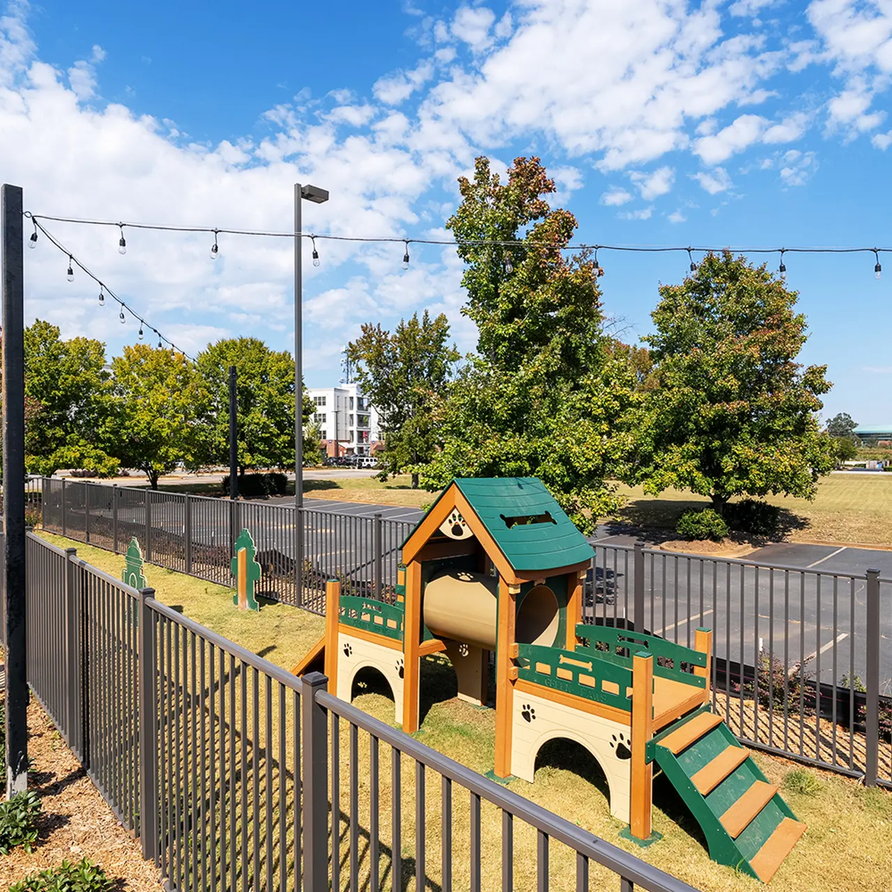 A colorful playground structure with slides and a tunnel, surrounded by a grassy area and trees. In the background, there are strings of lights and an open field with parked vehicles.