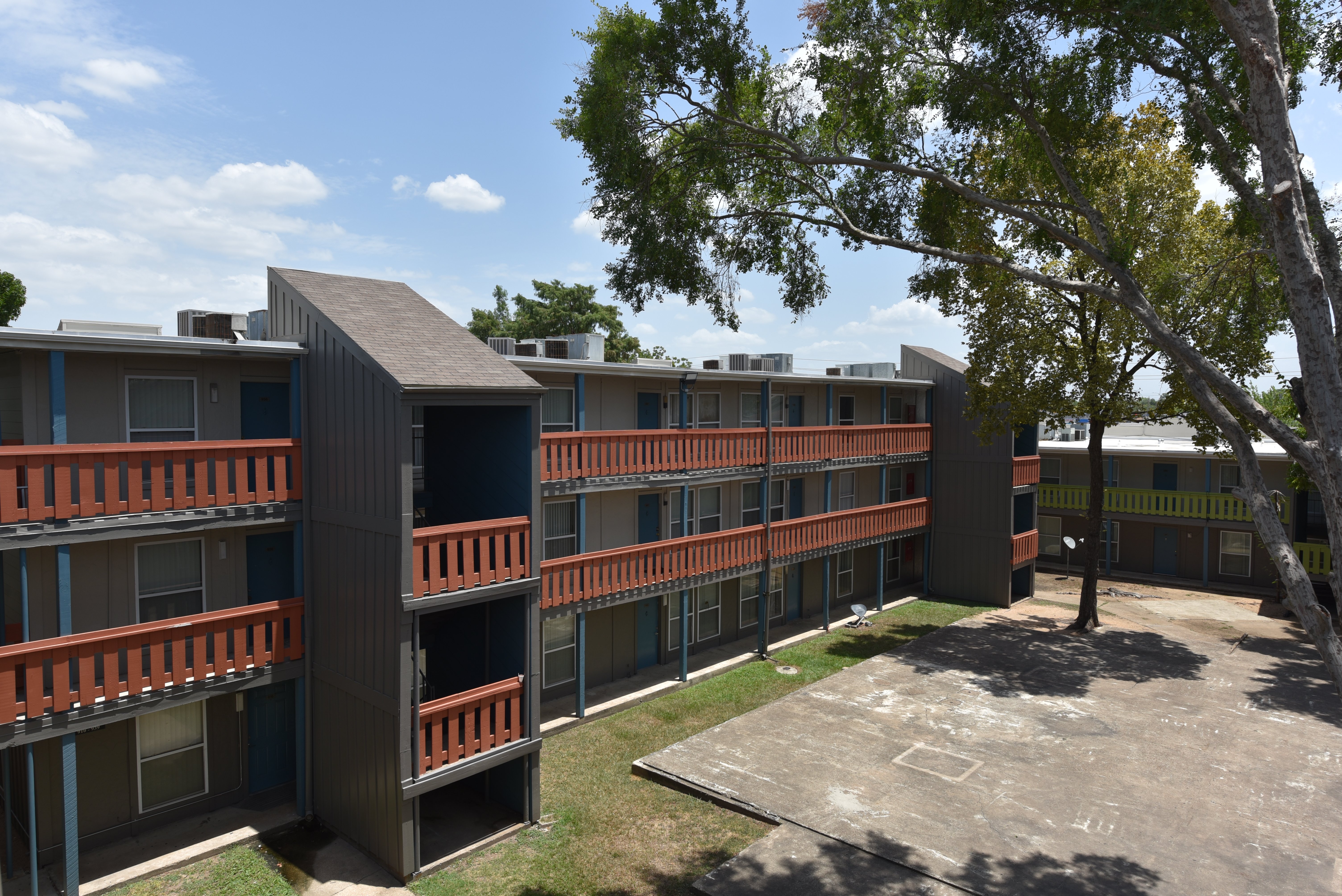 View of a multi-story apartment complex with balconies, surrounded by trees and blue sky.