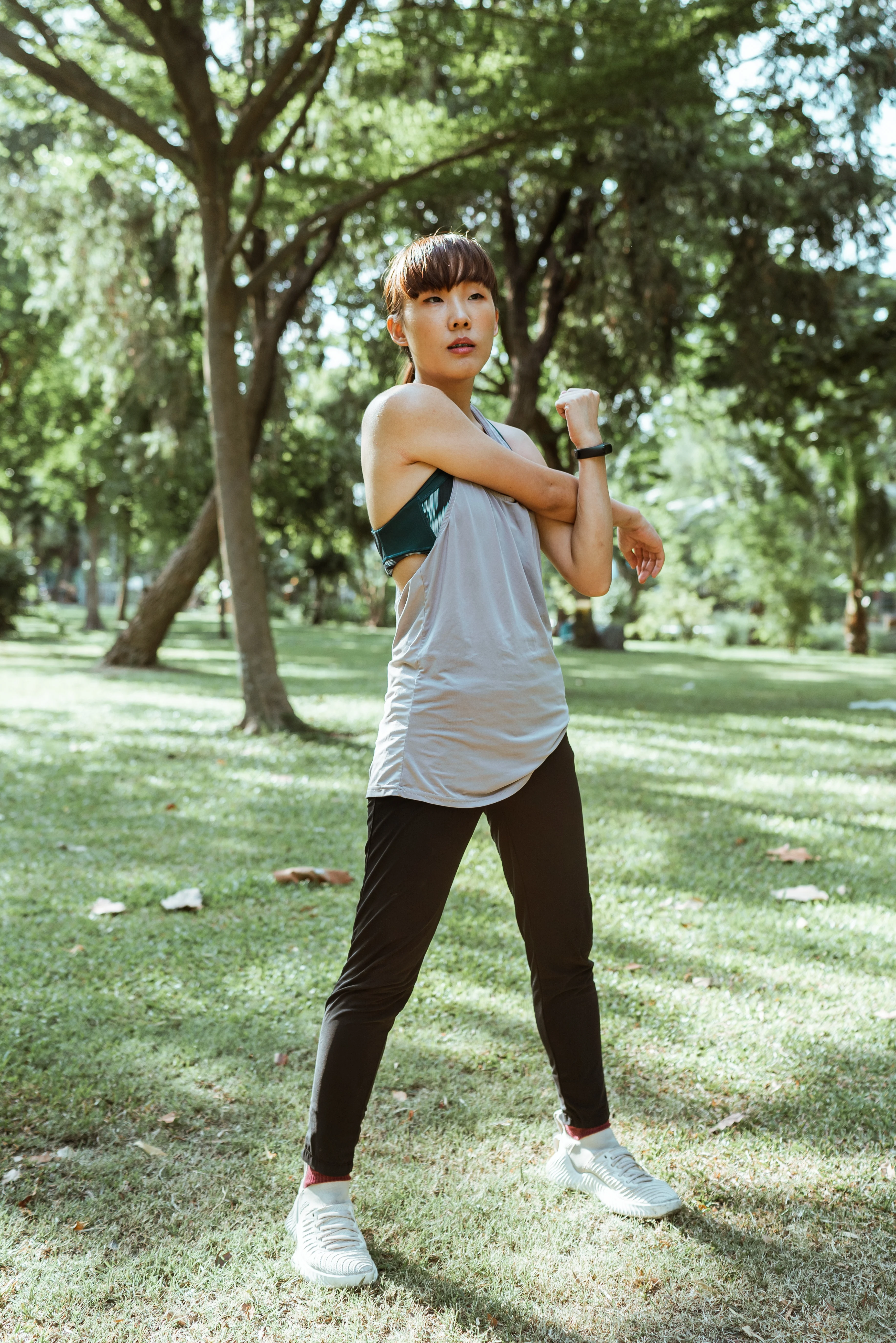 Woman Exercising in a Park A woman stretching her arm in a park, wearing a gray tank top and black leggings, with trees and grass in the background.