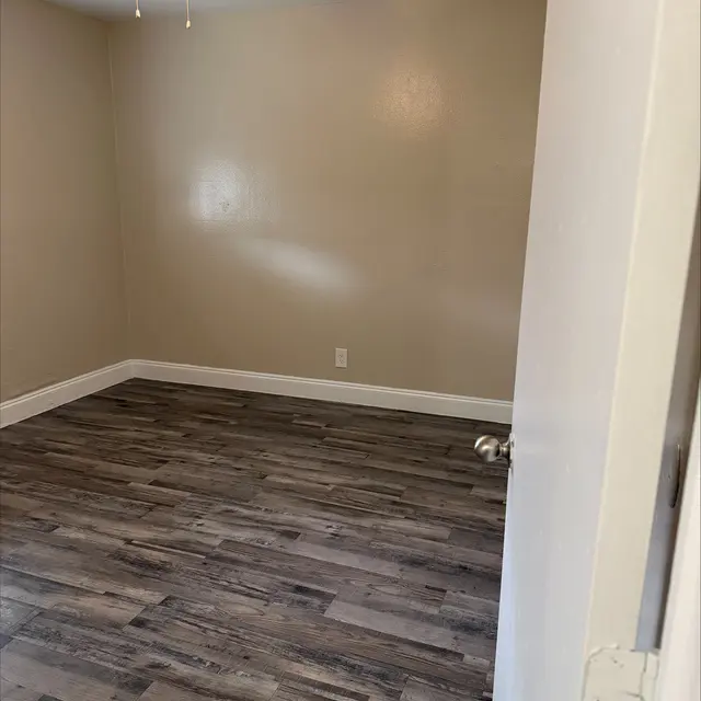 A clean, empty room with light-colored walls and a wooden floor. There is a ceiling fan visible on the ceiling, and the room is well-lit.