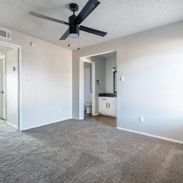 A cozy room featuring a ceiling fan, light-colored walls, and plush carpet flooring. One wall has a window letting in natural light, while another wall has doorways leading to adjacent rooms. There is a small countertop area visible in the background.