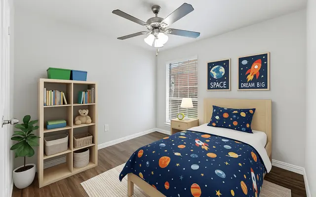 A bright children's bedroom featuring a bed with a space-themed comforter, a nightstand with a lamp, and a window with natural light. A bookshelf contains colorful books and toys, accompanied by a small potted plant in the corner.