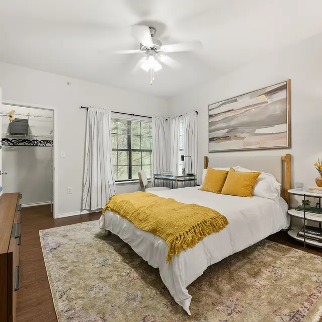 A well-decorated bedroom featuring a large bed with white linens and a yellow throw blanket, a dark wooden dresser, a floor lamp, and a colorful rug.