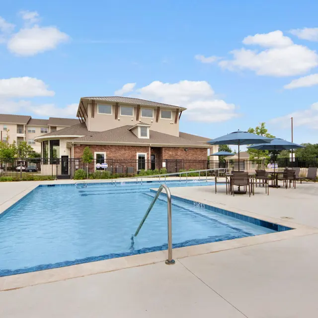 Swimming Pool Area at Apartment Complex A swimming pool area surrounded by lounge chairs, tables, and umbrellas, with a brick building in the background.