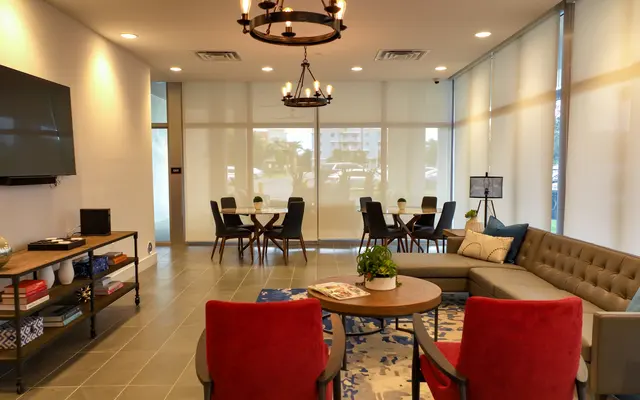 A modern lounge area featuring a sectional sofa, round tables, and chairs, illuminated by ceiling lights with a large TV mounted on one wall.