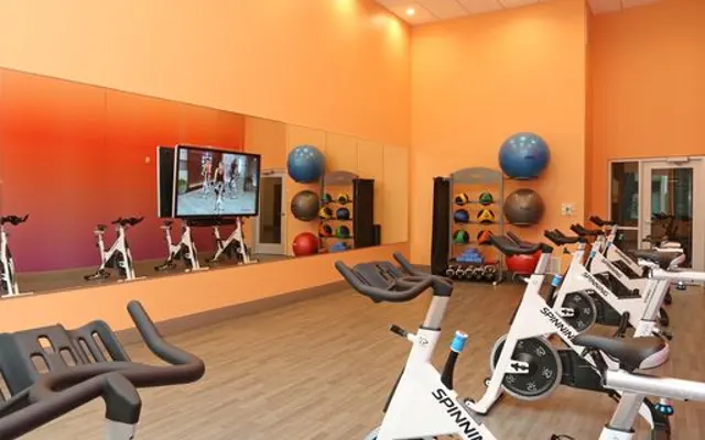 A brightly colored gym spinning room with exercise bikes, a large mirror, and a television showing a spinning class.