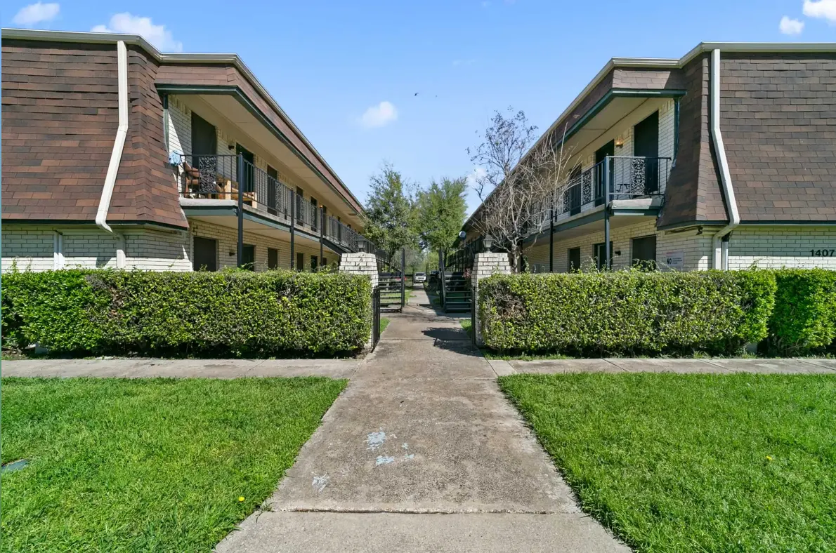 A view of an apartment complex with two blocks of buildings on either side of a concrete pathway. The buildings feature a brown and cream exterior, with balconies on the upper floors. Lush green bushes line the walkway, and there are a few trees in the background against a clear blue sky.