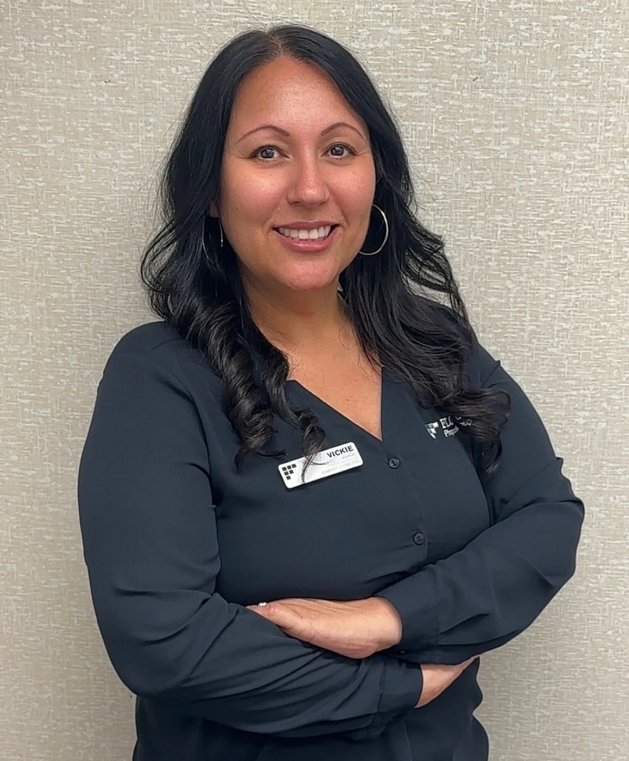 Professional Portrait A professional woman with long dark hair standing with arms crossed, wearing a black shirt and a name badge.