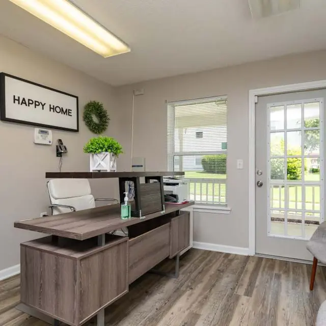 A cozy office space with a desk, chair, and greenery on the walls, featuring a large window and door leading to an outdoor area.