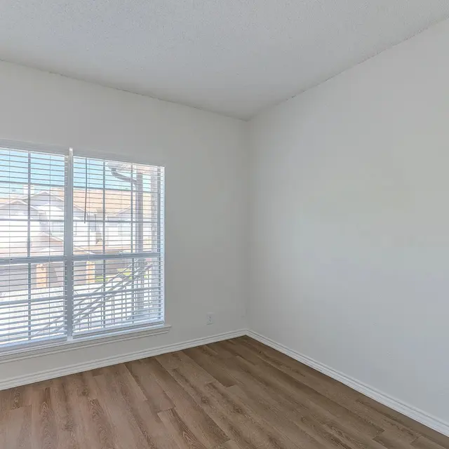 A bright, empty room with light-colored walls and wooden flooring. It features a large window with white blinds allowing natural light to fill the space.