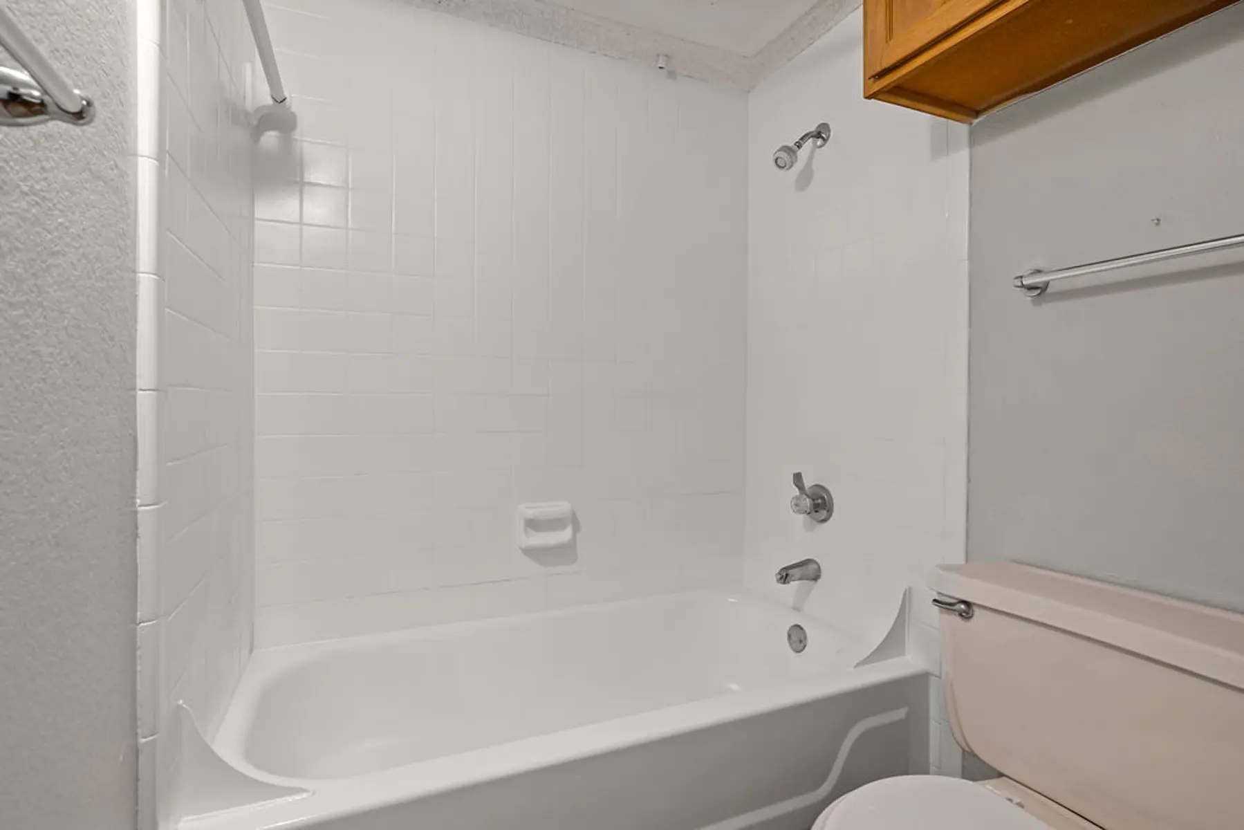 Simple Bathroom Interior A clean and simple bathroom featuring a white bathtub and shower combination, with light-colored tiles and a white toilet. There is a wooden cabinet above the bathtub.