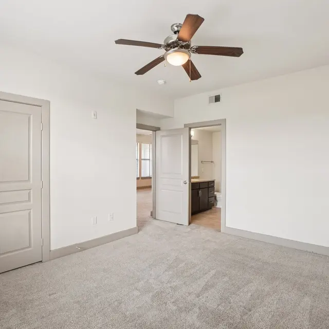 An empty bedroom featuring a ceiling fan, light-colored walls, and carpeted flooring. There are double doors on the left, leading to a closet, and an open door on the right that connects to another room, possibly a bathroom.