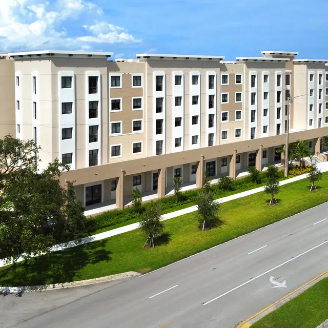 A modern four-story apartment building with a landscaped area and a road in front.