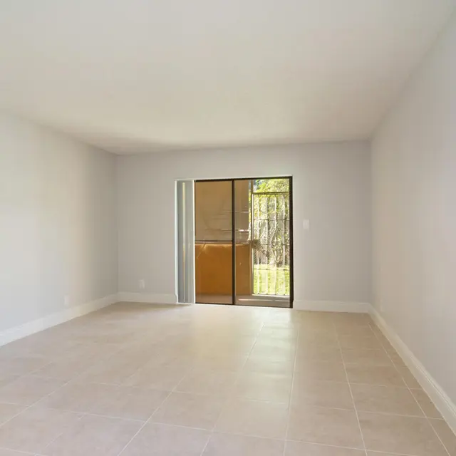 A spacious, empty room with light gray walls and beige tiled floor. A sliding glass door provides access to a small outdoor area. The room has a modern aesthetic with natural light streaming in through the door.