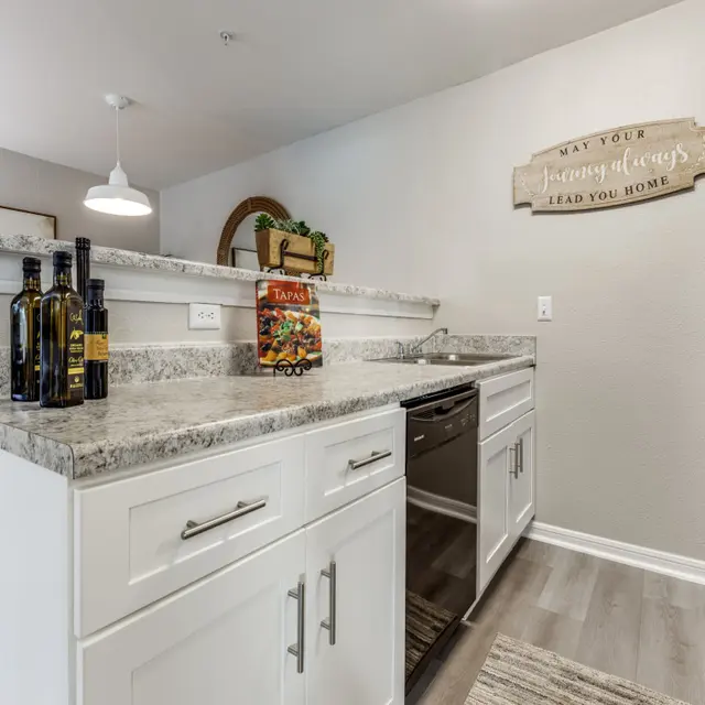 A stylish modern kitchen featuring white cabinets, granite countertops, and stainless steel appliances. A small wall shelf holds cooking oils and a colorful recipe book, with decorative items adding charm to the space.