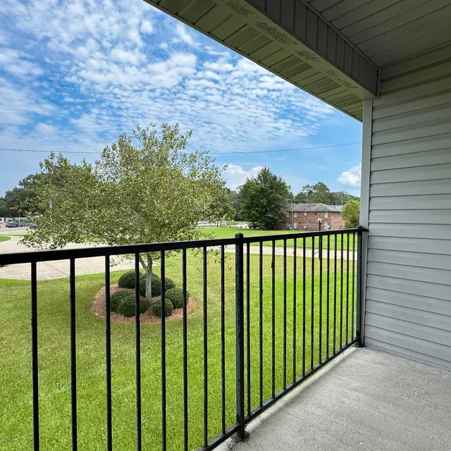 View from a balcony overlooking a green lawn with a tree and a sidewalk in the distance under a partly cloudy sky.