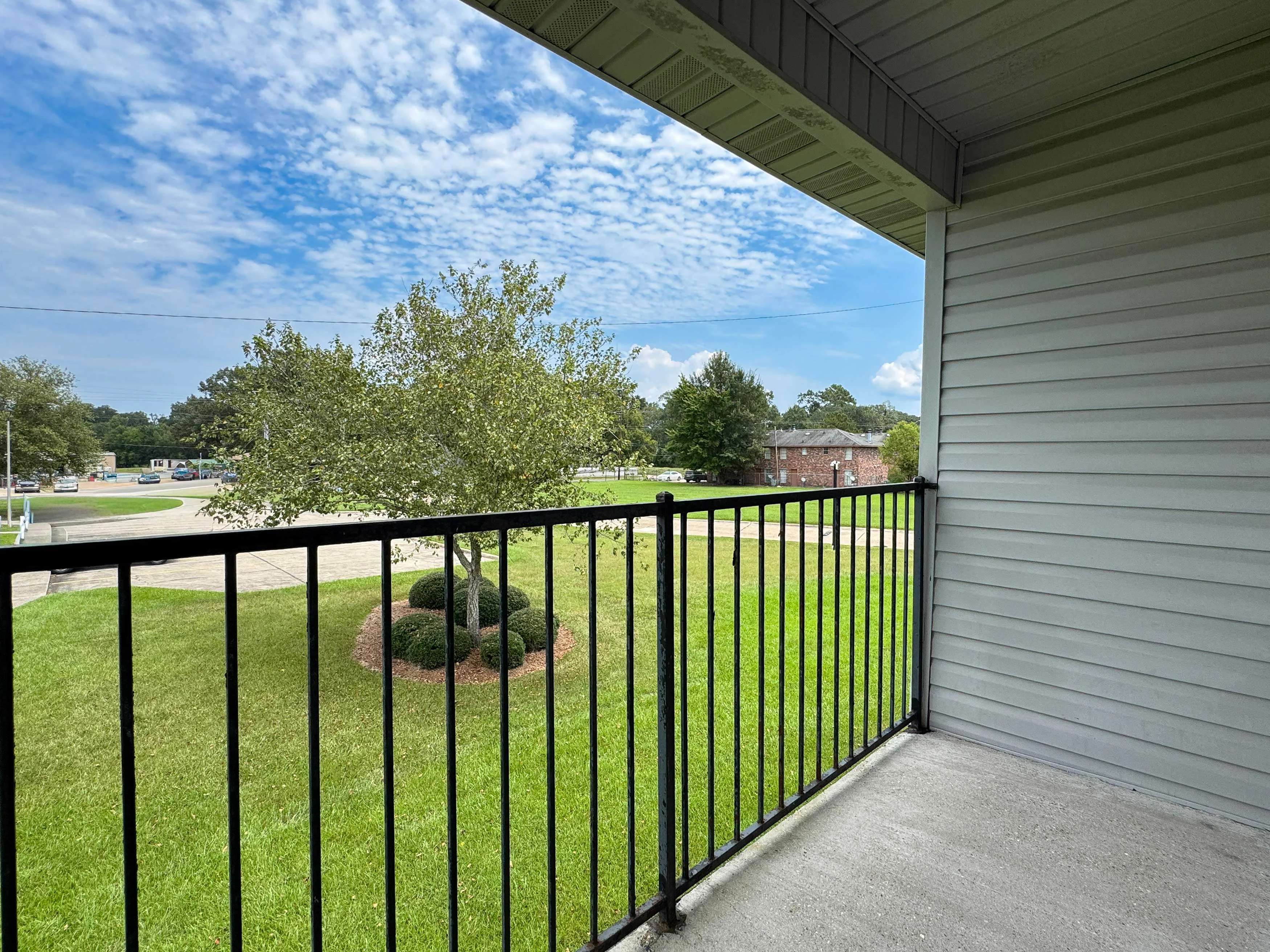 View from a balcony overlooking a green lawn with a tree and a sidewalk in the distance under a partly cloudy sky.